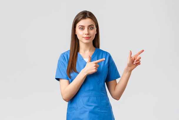 Healthcare workers, prevent virus, insurance and medicine concept. Young nurse or doctor in blue scrubs pointing fingers right, recommend banner or promo for clinic patients - Foto, imagen
