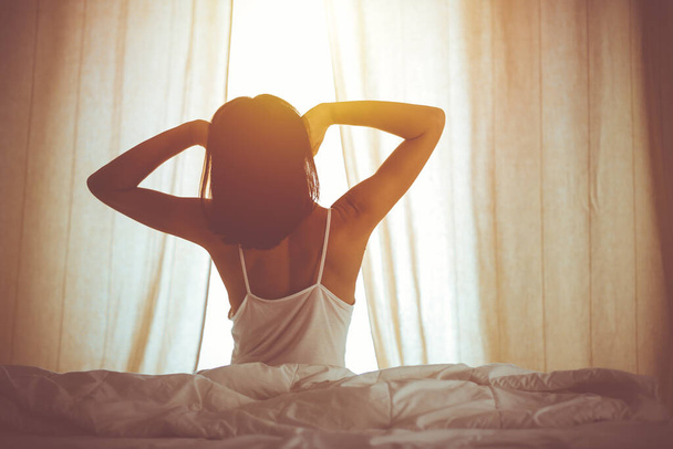 Woman stretching hands in bed after wake up, sun flare . Brunette entering a day happy and relaxed after good night sleep and back view. Concept of a new day and joyful weekend - Foto, Imagen