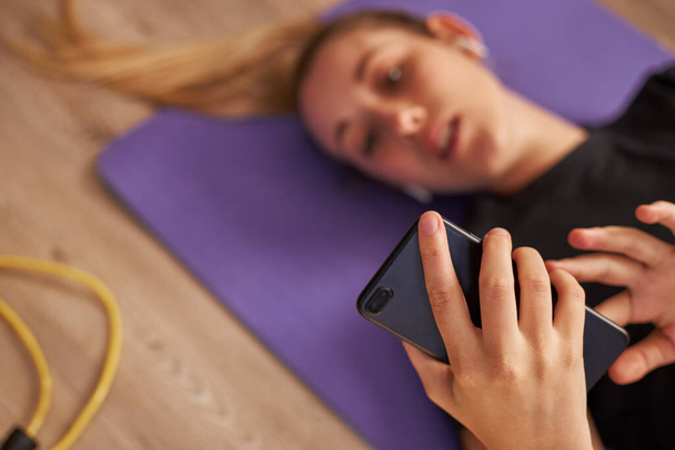 Mladá žena ležící na podložce jógy na mobilním telefonu a poslouchající hudbu. - Fotografie, Obrázek