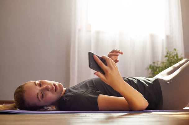 Young woman lying on yoga mat on mobile phone listening to music. - Photo, Image