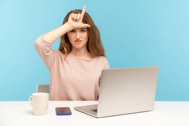 Lost job, unemployment. Depressed young woman employee sitting at workplace with laptop and showing loser gesture, desperate about unlucky dismissal. indoor studio shot isolated on blue background - Φωτογραφία, εικόνα