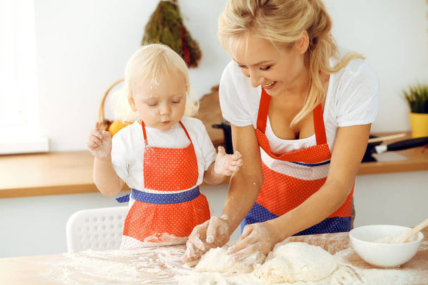 Little girl and her blonde mom in red aprons playing and laughing while kneading the dough in kitchen. Homemade pastry for bread, pizza or bake cookies. Family fun and cooking concept - 写真・画像