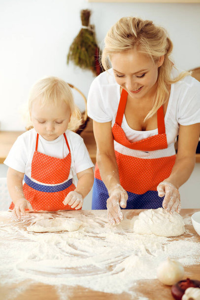 Little girl and her blonde mom in red aprons playing and laughing while kneading the dough in kitchen. Homemade pastry for bread, pizza or bake cookies. Family fun and cooking concept - Foto, imagen