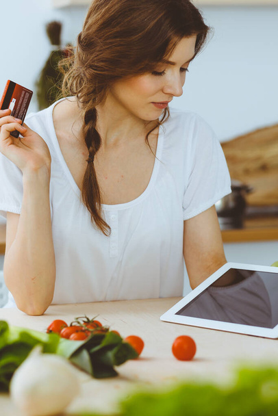 Young woman looking for a new recipe for cooking in a kitchen. Housewife is making online shopping by tablet computer and credit card - Foto, Imagem