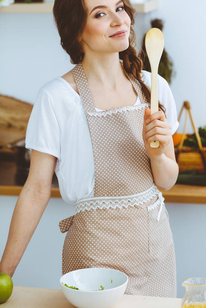 Young brunette woman cooking in kitchen. Housewife holding wooden spoon in her hand. Food and health concept - Fotografie, Obrázek