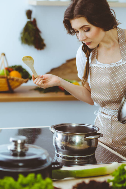 Young brunette woman cooking soup in kitchen. Housewife holding wooden spoon in her hand. Food and health concept - Foto, imagen
