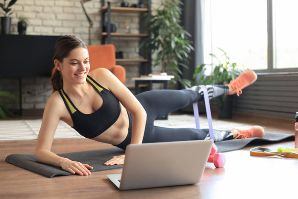 Fitness beautiful slim woman doing side plank with an elastic band and watching online tutorials on laptop, training in living room. Stay at home activities. - Photo, image