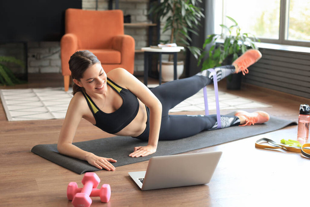 Fitness beautiful slim woman doing side plank with an elastic band and watching online tutorials on laptop, training in living room. Stay at home activities. - Photo, image