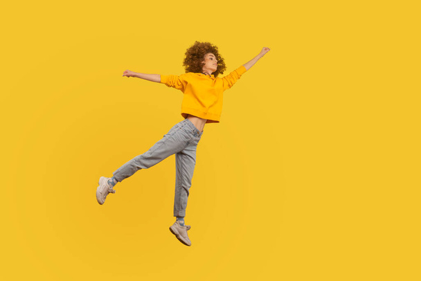 Portrait of superhero curly-haired girl in urban style outfit flying with raised hand high in air, feeling superpower and inspiration, achieving goal. indoor studio shot isolated on yellow background - Foto, afbeelding