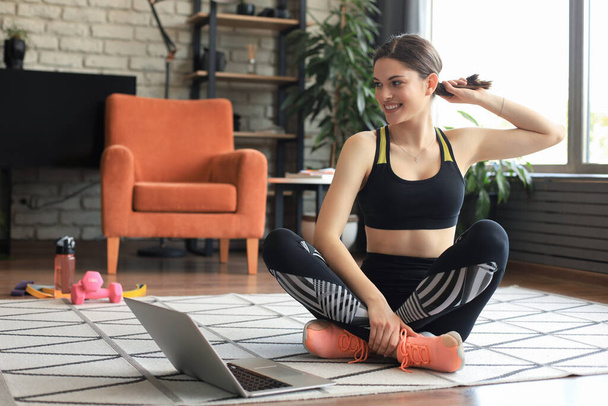 Fitness beautiful slim woman is sitting on the floor with dumbbells and bottle of water using laptop at home in the living room. Stay at home activities - Foto, Imagem