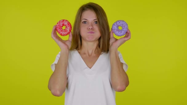 Woman looks at a delicious blue, red donuts in hands with pouting cheeks, mouth - Footage, Video