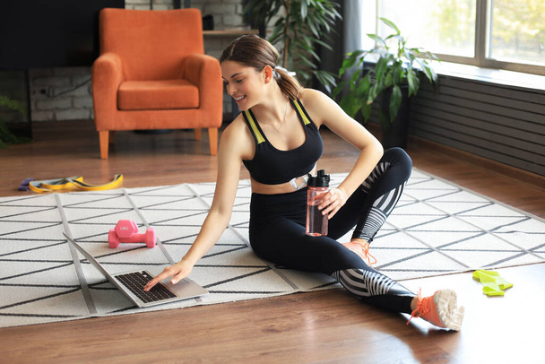 Fitness beautiful slim woman is sitting on the floor with dumbbells and bottle of water using laptop at home in the living room. Stay at home activities - Фото, зображення