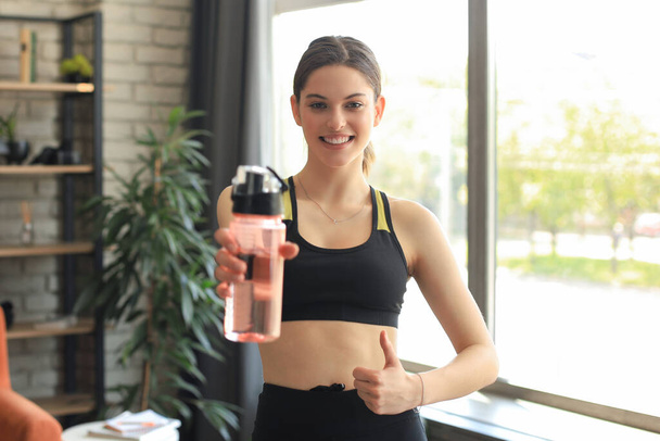 Young beautiful woman standing with dumbbell and drink water after exercising looking camera. Attractive female bodybuilder working out. Fitness and healthy lifestyle concept - Foto, Bild