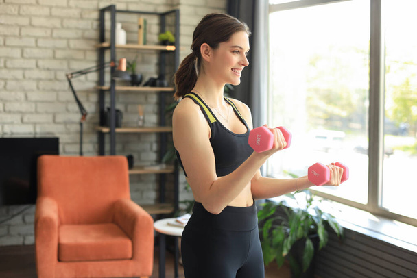 Young beautiful sports girl in leggings and a top does exercises with dumbbells. Healthy lifestyle. A woman goes in for sports at home - Foto, afbeelding