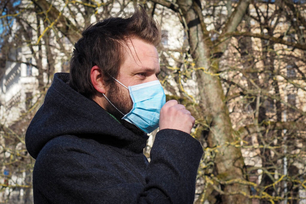 Красивый молодой европеец кашляет на улице в медицинской маске. Крупный план 35-летнего мужчины в респираторе для защиты от инфекции гриппом или коронавирусом (Covid-19
) - Фото, изображение