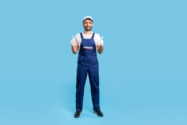 Full length workman in overalls, cap and protective gloves standing, showing thumbs up, like gesture and smiling, recommending good house repair services. studio shot isolated on blue background - Foto, afbeelding
