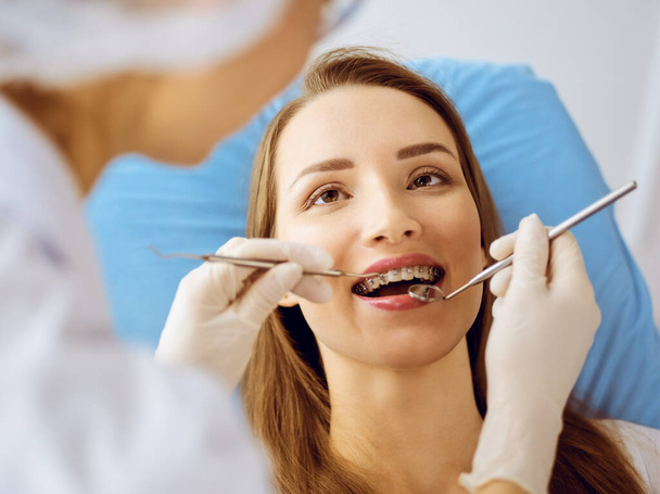 Mujer joven sonriente con corchetes de ortodoncia examinados por el dentista en la clínica dental. Concepto de dientes sanos y medicina - Foto, Imagen