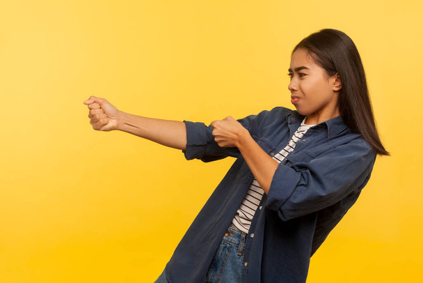 Portrait of diligent purposeful girl in denim shirt pulling invisible heavy burden, striving hard to achieve success, dragging with persistence. indoor studio shot isolated on yellow background - Zdjęcie, obraz