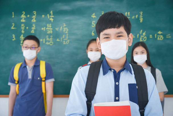 Jonge aziatische schooljongen dragen gezichtsmasker staan voor schoolbord en kijken naar de camera, klasgenoot staan op de achtergrond. - Foto, afbeelding