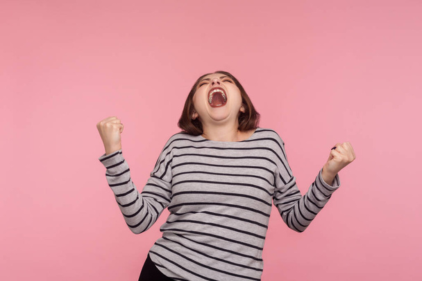 Triumphal victory! Portrait of extremely happy, overjoyed woman in striped sweatshirt yelling for joy with raised fists, celebrating success, euphoria from win. studio shot isolated on pink background - Fotografie, Obrázek