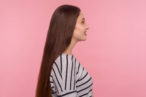 Profile of joyful carefree young woman in striped sweatshirt smiling to side copy space, satisfied and rejoicing happy healthy life, optimistic outlook. indoor studio shot isolated on pink background - Fotó, kép