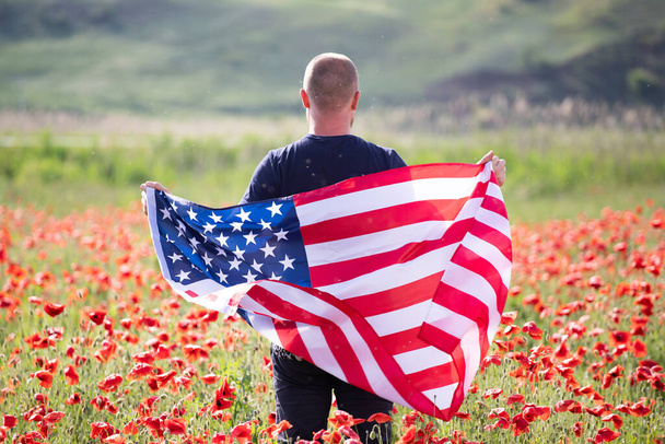 Attractive man holding Flag of the United States in beautiful poppy field on a clear, sunny day. Celebrating Independence Day, National holiday concept. - Фото, изображение