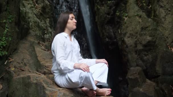 woman meditates in lotus position on rock against waterfall - Footage, Video