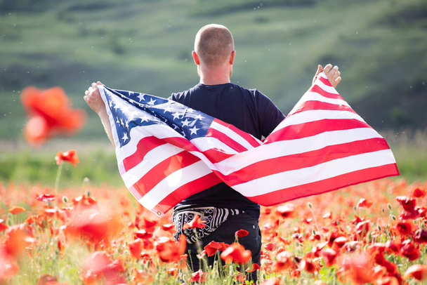 Attractive man holding Flag of the United States in beautiful poppy field on a clear, sunny day. Celebrating Independence Day, National holiday concept. - Photo, image