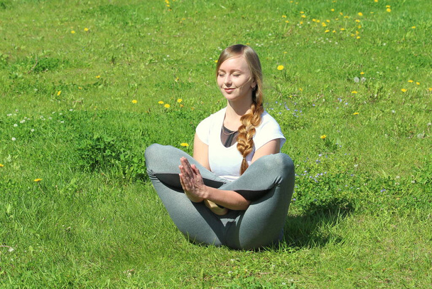 Young woman of European appearance does yoga in summer nature. Woman sitting in flowering lotus pose, vikasitakamalasana. High quality photo for web and print with empty space for text and design. - Фото, изображение