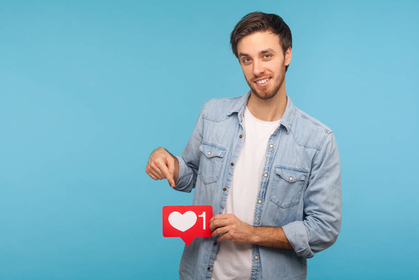 Click Like and subscribe! Portrait of handsome blogger man in denim shirt pointing at social media heart button, emoji counter, follower notification. indoor studio shot isolated on blue background - Фото, изображение