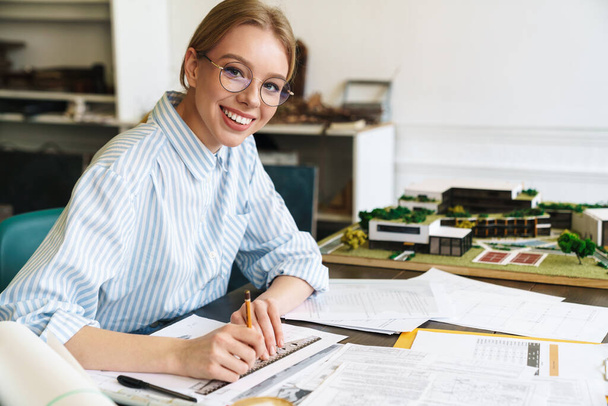 Foto de una mujer rubia sonriente arquitecta con anteojos que trabaja con dibujos mientras diseña borrador en el lugar de trabajo
 - Foto, Imagen