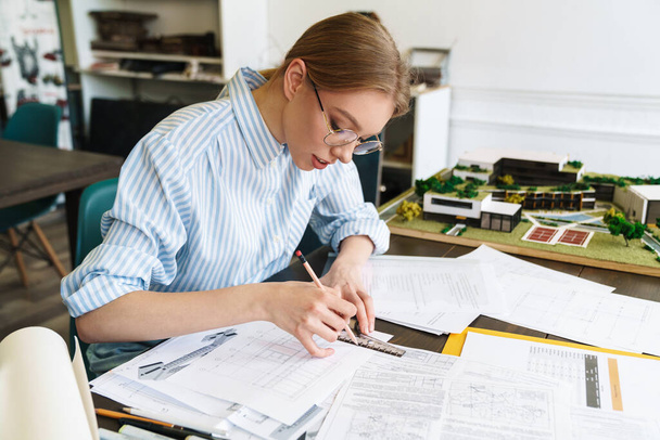 İş yerinde taslak tasarlarken gözlüklü, çizimlerle uğraşan odaklanmış genç kadın mimarın fotoğrafı - Fotoğraf, Görsel
