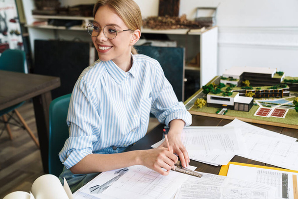 Foto de una mujer rubia sonriente arquitecta con anteojos que trabaja con dibujos mientras diseña borrador en el lugar de trabajo
 - Foto, imagen