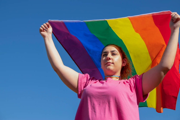 Mujer joven ondeando la bandera del arco iris con una camisa rosa y un collar de arco iris en un cielo azul claro. Concepto de Lgtbiq
. - Foto, imagen