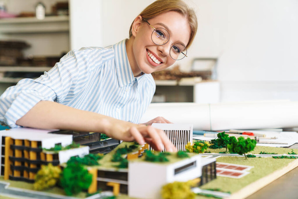 Photo of joyful young woman architect in eyeglasses designing draft with house model and sitting at workplace - Foto, imagen