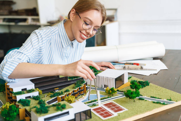 Photo of joyful young woman architect in eyeglasses designing draft with house model and sitting at workplace - Fotoğraf, Görsel