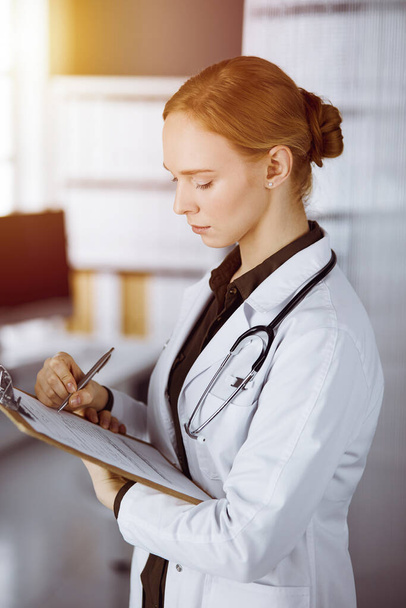 Cheerful smiling female doctor using clipboard in sunny clinic. Portrait of friendly physician woman at work. Medicine concept - Foto, imagen