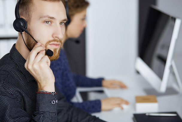 Red-bearded businessman talking by headset near his female colleague while sitting in modern office. Diverse people group in call center. Telemarketing and customer service - Fotó, kép