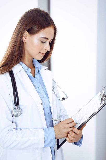 Doctor woman filling up medical form while standing near window in clinic. Physician at work. Medicine and health care concept - Photo, Image