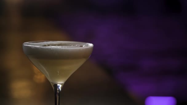 Primer plano de bebida alcohólica con espuma en vaso de martini. Imágenes de archivo. Un cóctel de color amarillo de pie sobre fondo púrpura y marrón de una habitación oscura
. - Metraje, vídeo