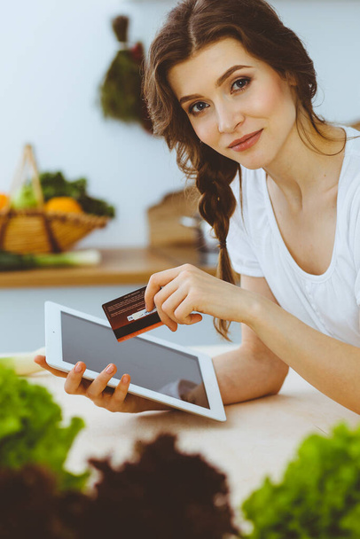 Young woman looking for a new recipe for cooking in a kitchen. Housewife is making online shopping by tablet computer and credit card - Valokuva, kuva