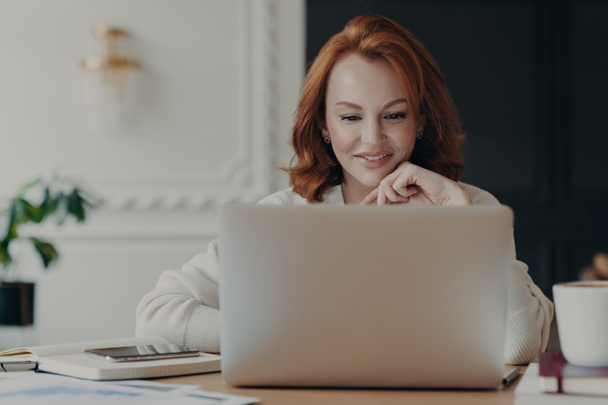 Опытная рыжеволосая деловая женщина читает публикацию на современном ноутбуке, концентрируется на экране с приятной улыбкой, проводит исследования для планирования запуска, проверяет почтовый ящик, работает из дома
 - Фото, изображение