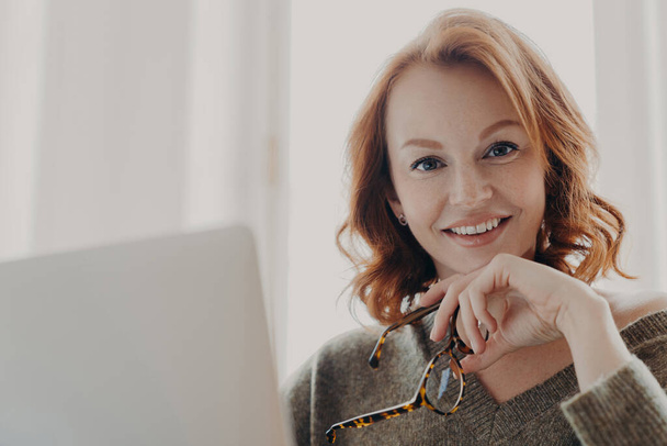 きれいな生姜のビジネスの女性は、快適な笑顔を持っている、ノートパソコンのコンピュータを介してオンラインのウェブ情報を検索光メガネを保持し、同僚のデスクトップに座って、フリーランスの仕事で忙しい。技術コンセプト - 写真・画像