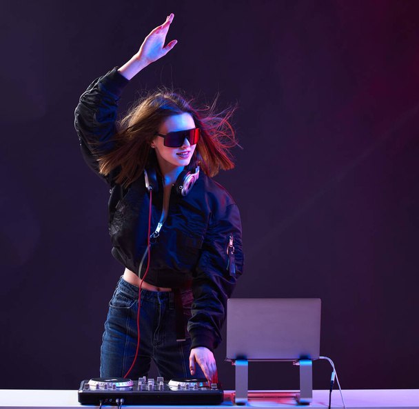 Stylish DJ girl with headphones and glasses on the bomber jacket mixes the music at the party and neon lights. - Foto, Bild