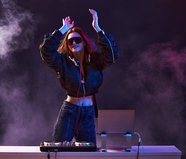 Stylish girl DJ in glasses headphones and a bomber mixes music dancing at a party in the neon light in the smoke . - 写真・画像