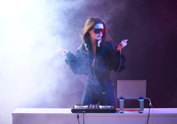 Stylish girl DJ in glasses headphones and a bomber mixes music dancing at a party in the neon light in the smoke . - Фото, изображение