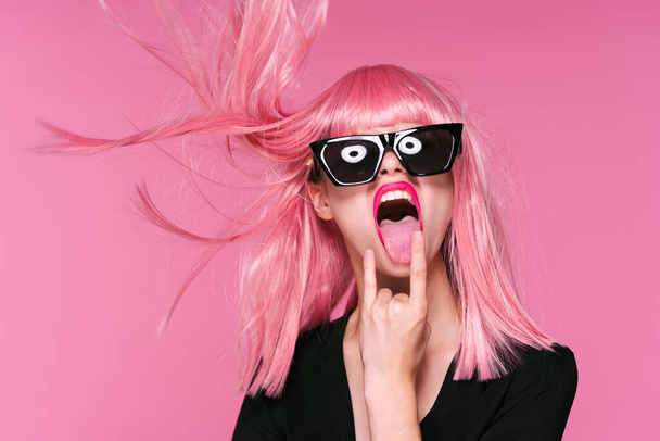 Νεαρή γυναίκα με ροζ περούκα που δείχνει γλώσσα σε ροζ φόντο - Φωτογραφία, εικόνα
