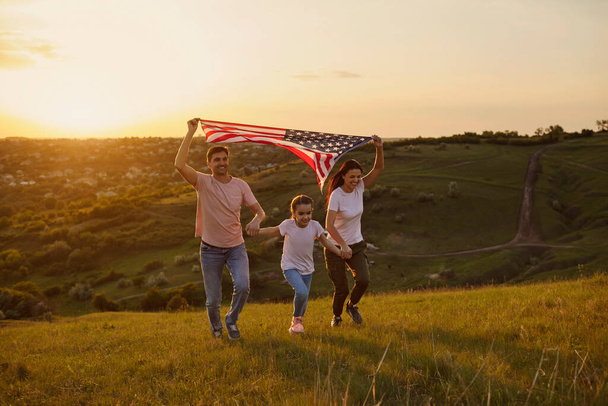 Szczęśliwa rodzina z flagą usa biegnie po przyrodzie wieczorem o zachodzie słońca. Matka ojciec i córka świętują zabawę Dzień Niepodległości Ameryki. - Zdjęcie, obraz