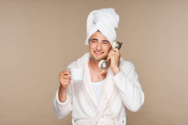 Young emotional man in bathrobe with cup of coffee talking on vintage phone. Studio shot. Isolated - Фото, изображение