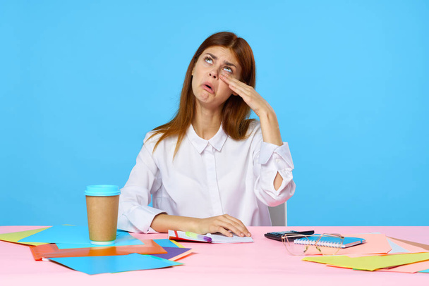 jonge zakenvrouw zitten aan bureau en huilen geïsoleerd op blauwe achtergrond  - Foto, afbeelding